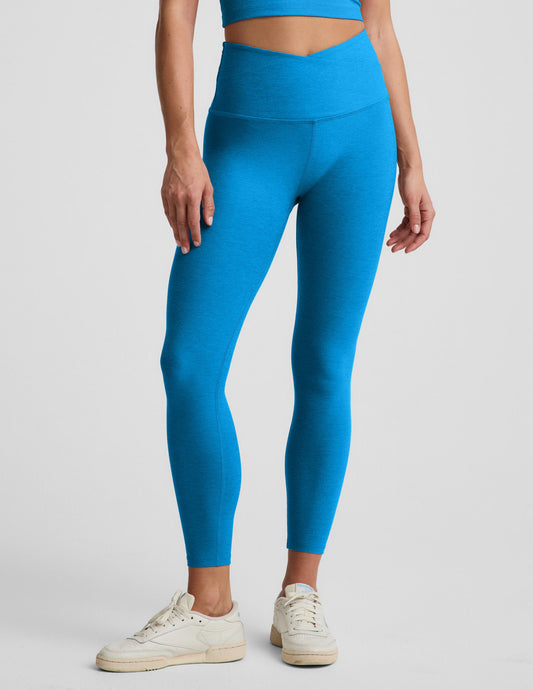 Woman wearing a blue cropped tank top and blue high waisted midi leggings with a v front waistband against a plain background.