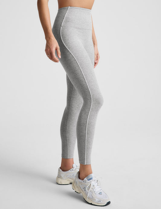 Woman wearing a gray sports bra and leggings with white contrast piping on a plain background.
