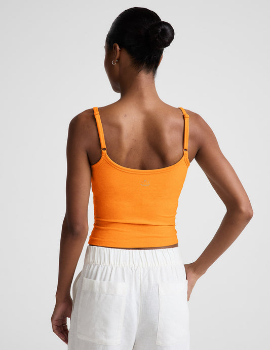 Woman wearing an orange tank top and white linen pants against a plain background.