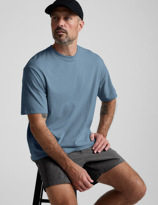 A man sitting on a stool wearing the gray Saturday Men's Short and a blue t-shirt. The shorts have a mid-thigh length.