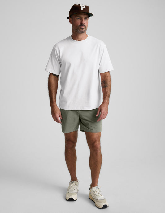 Man wearing olive Saturday Men's Short with a white t-shirt and brown cap, standing against a light grey background.