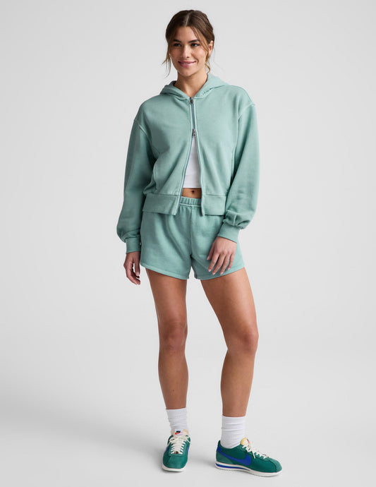 Woman wearing a green zip-up sweatshirt and green shorts against a plain background.
