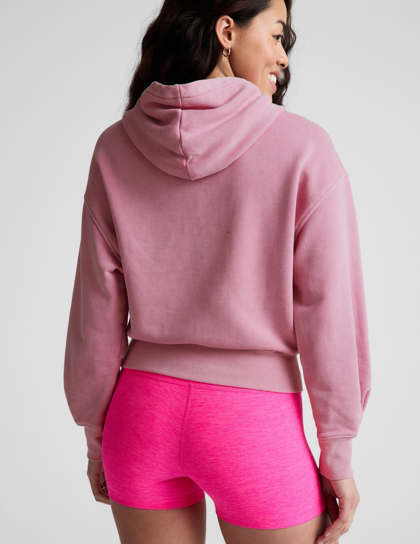 Woman wearing a pink sweatshirt and bright pink shorts on a plain background.