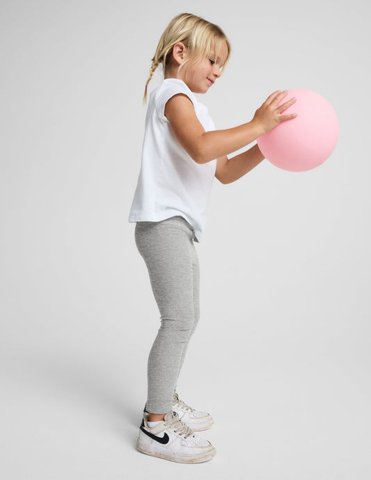 A young girl with blonde braids stands against a light gray studio background wearing a white short-sleeve t-shirt, light gray heathered leggings, and white sneakers.