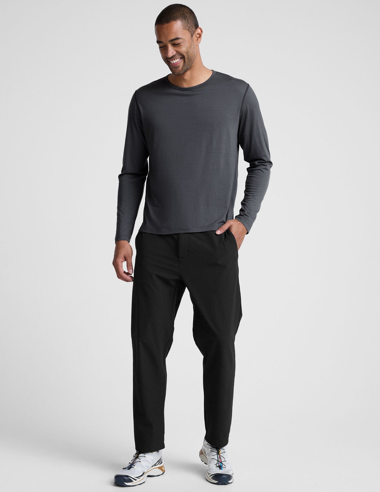Man wearing a dark gray crew neck long sleeve and black pants against a light background.