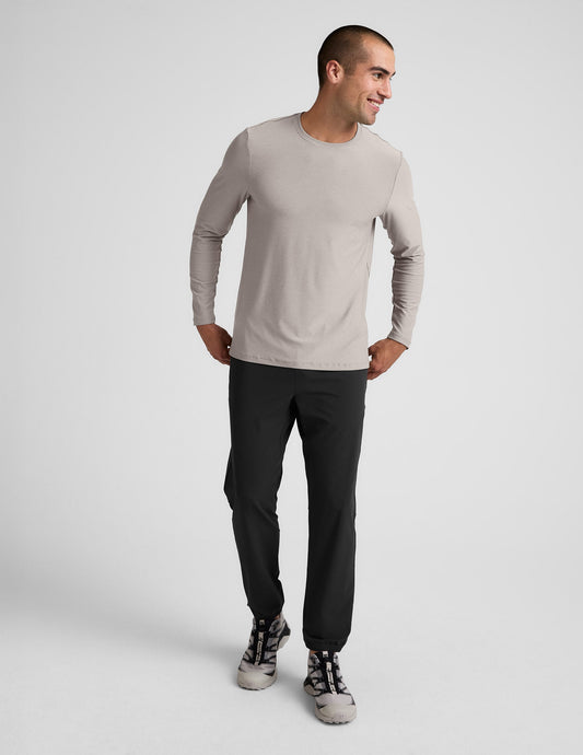 Man wearing a beige long-sleeve shirt and black pants against a plain background.
