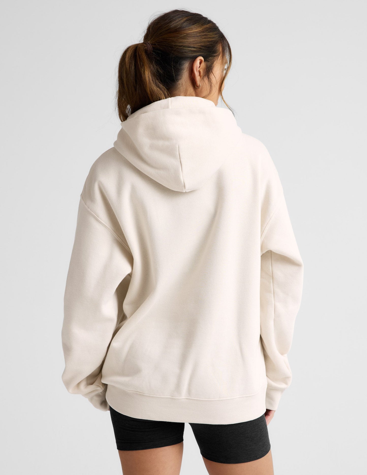Woman wearing a white oversized hoodie and black shorts on a light gray background.