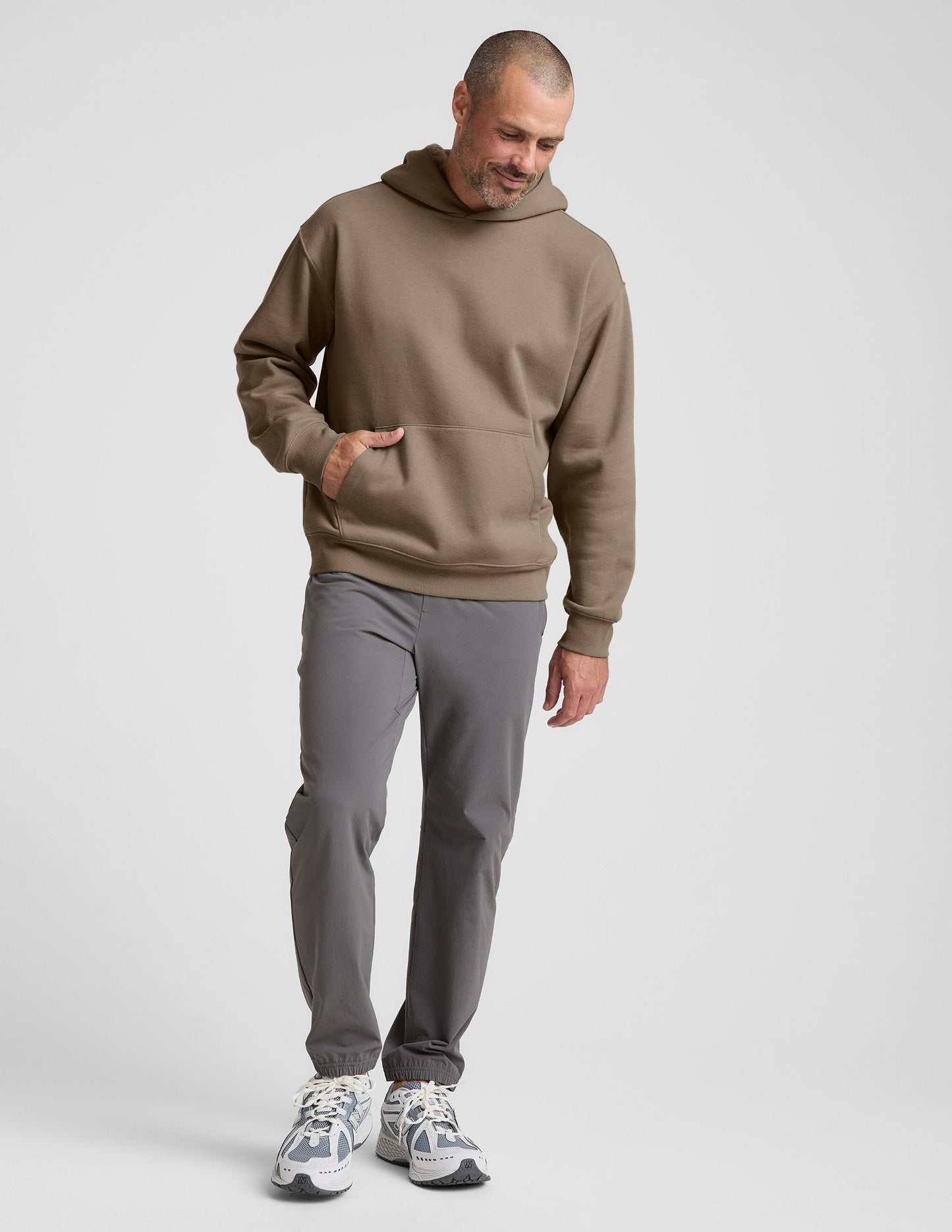 Man wearing a brown hoodie and gray pants on a plain background.