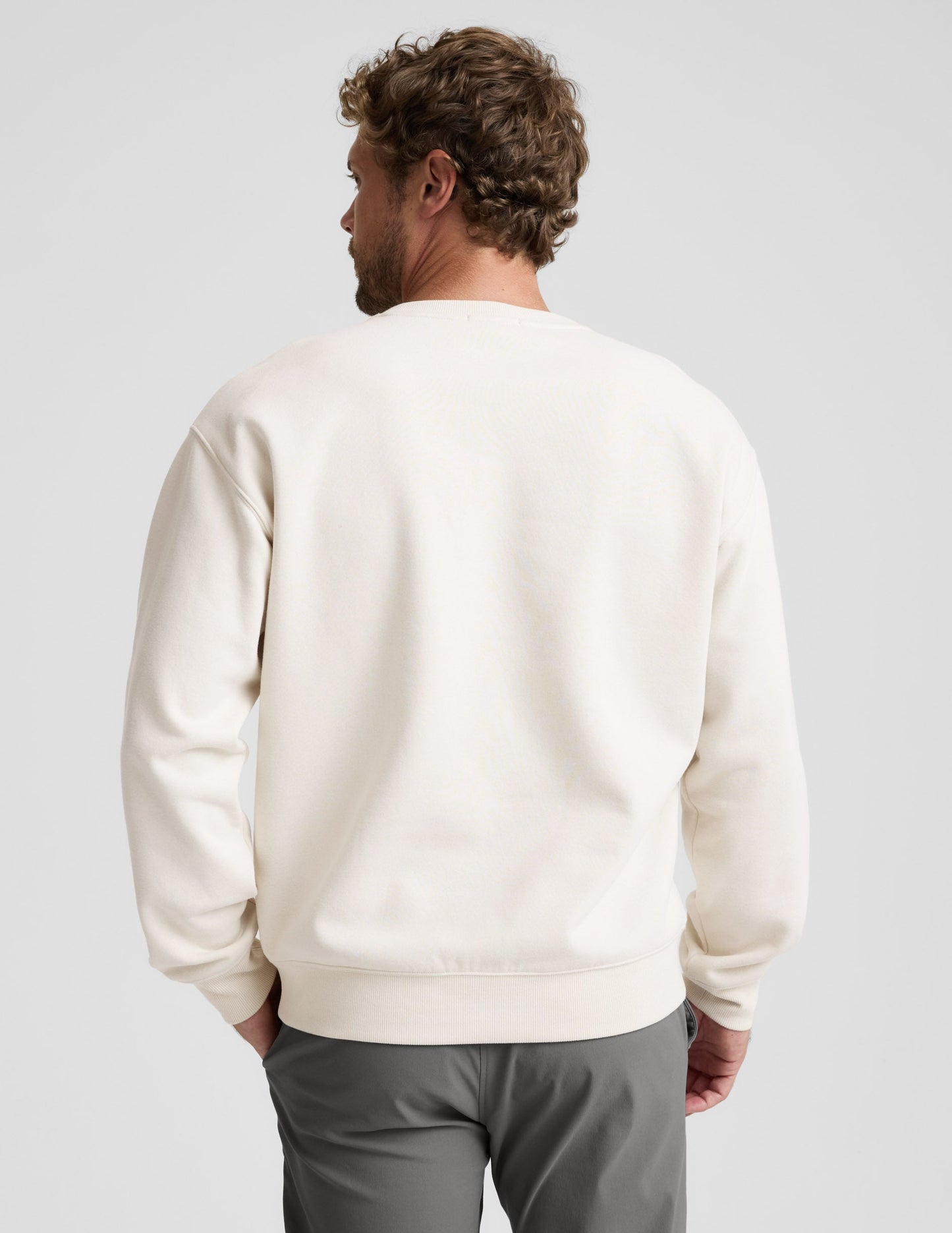 Man wearing a beige sweater and gray pants on a plain background.