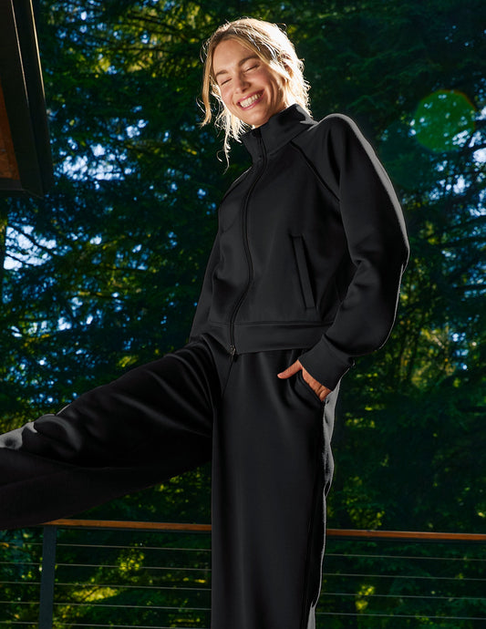 Woman in black jacket and pants standing outdoors with a blurred green background.