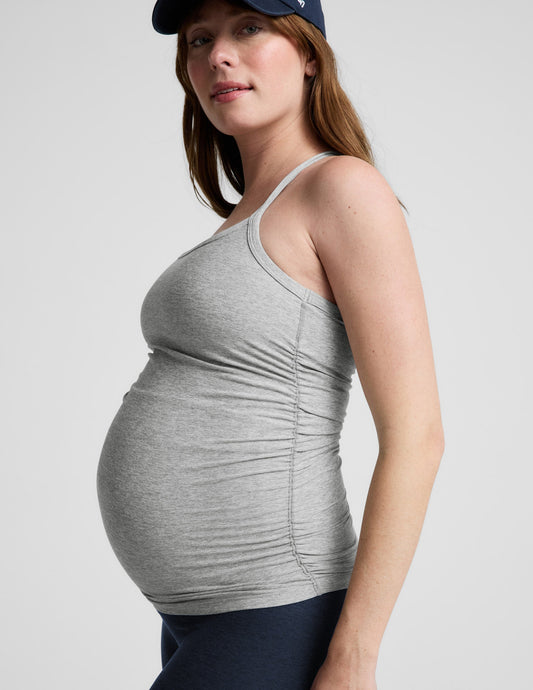 Woman wearing a gray maternity tank top and navy cap on a light gray background.