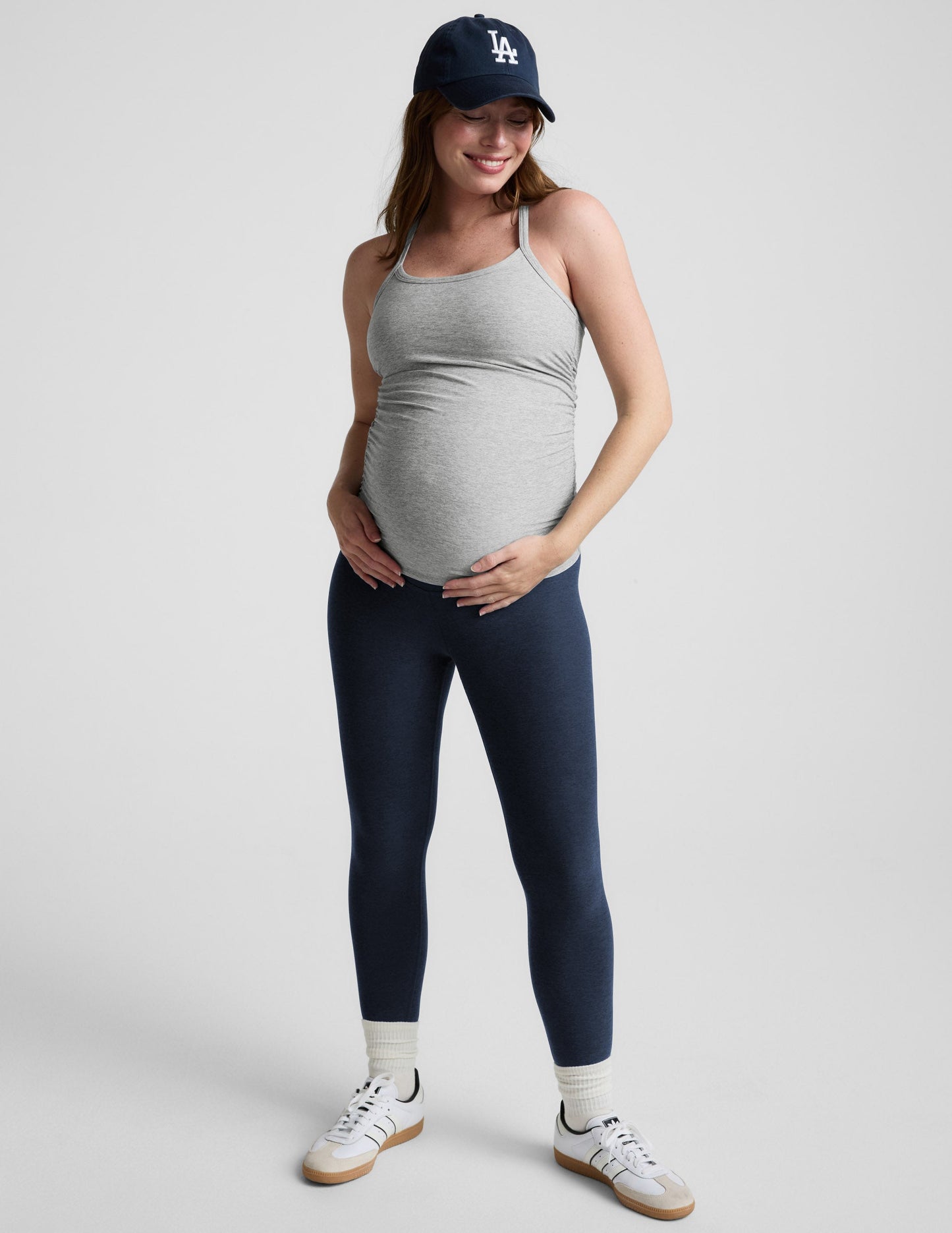 Woman wearing a gray maternity tank top and navy cap on a light gray background.