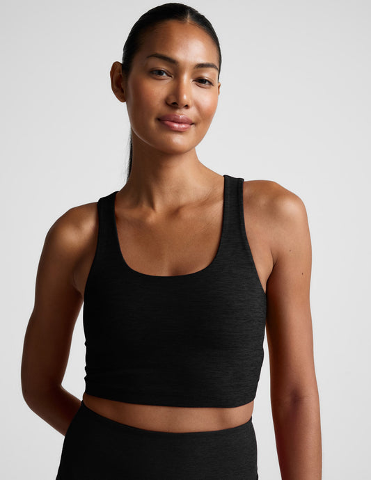 Woman in a black cropped tank with a crossover strap in the back and black leggings against a light gray background.