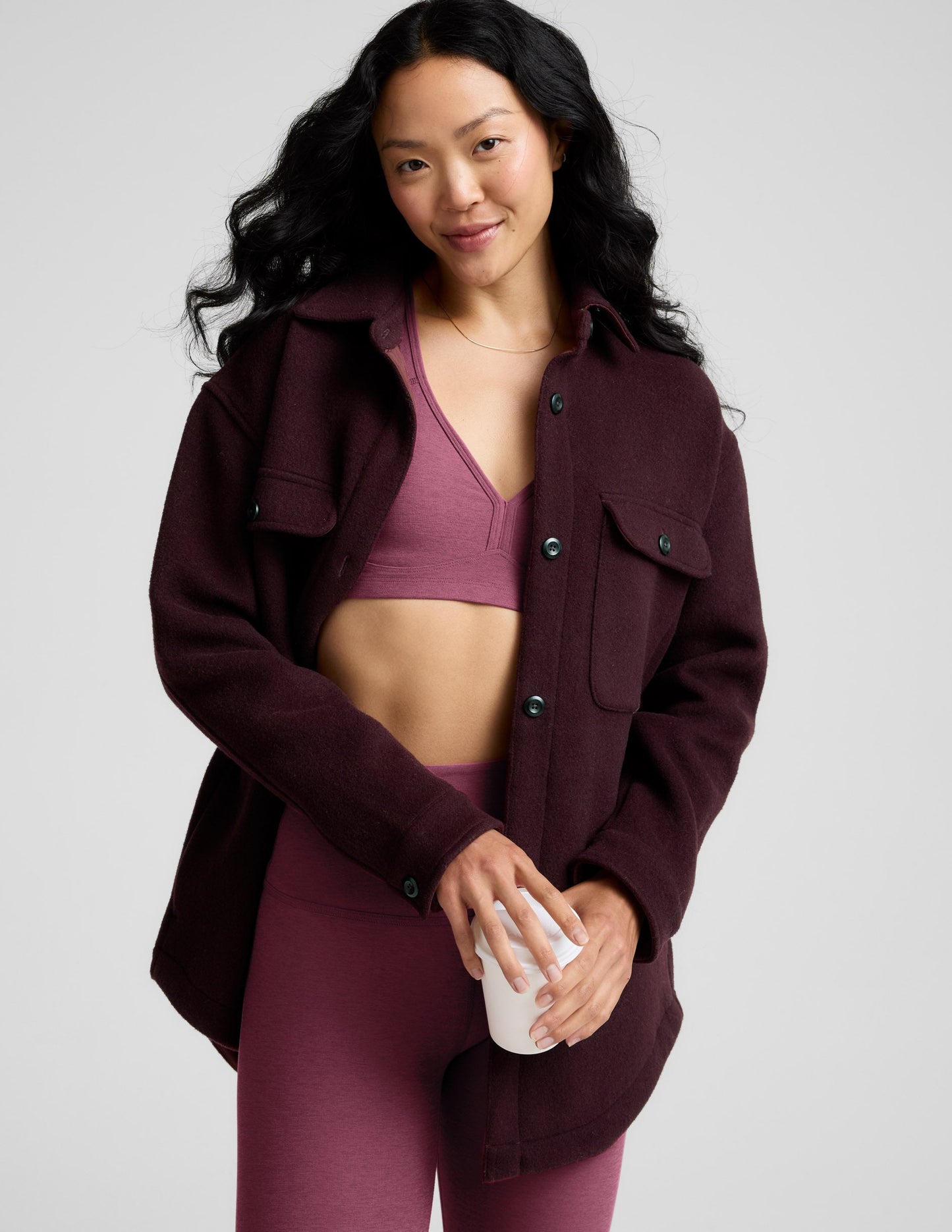 Woman wearing a maroon jacket over a sports bra and leggings, holding a white cup.