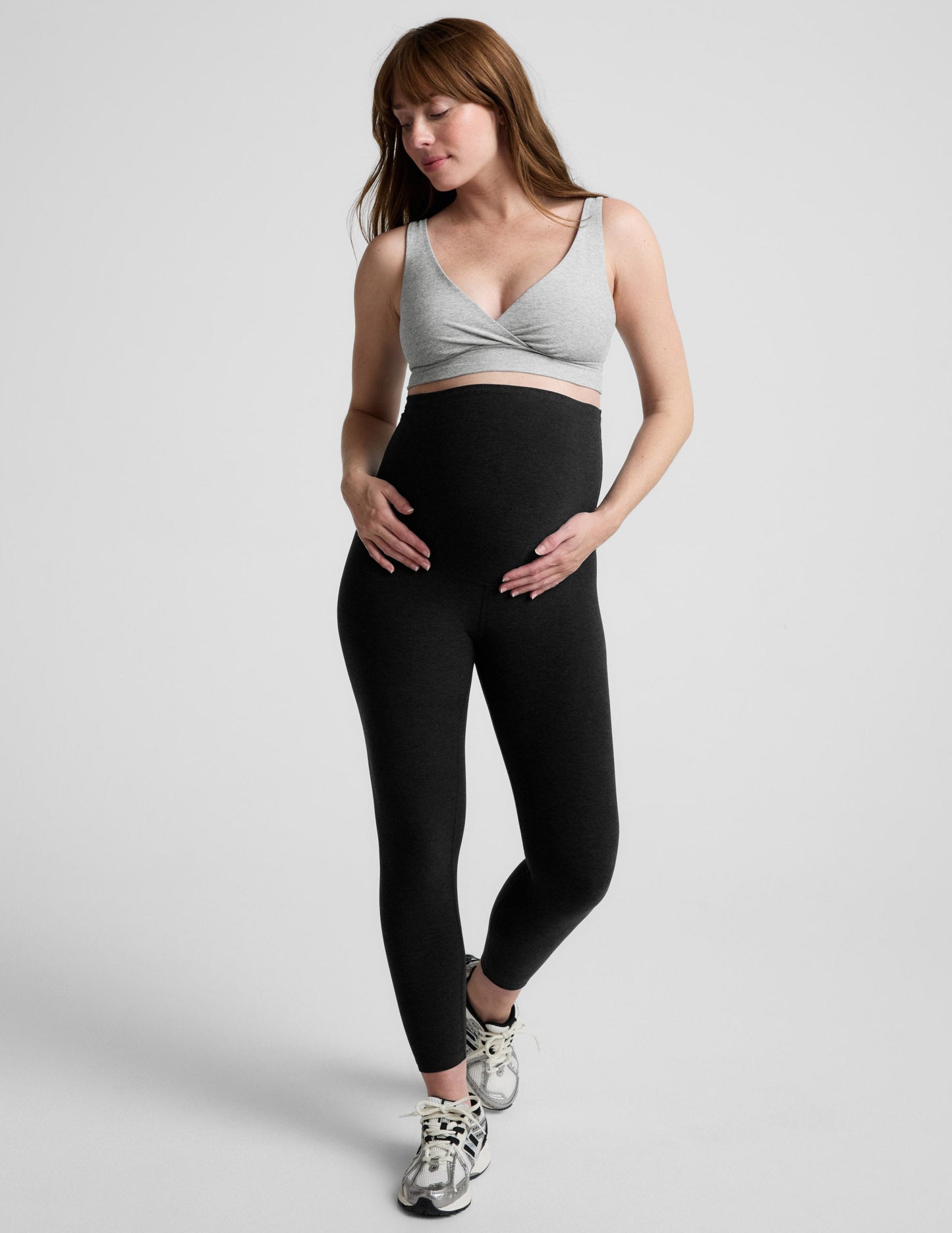 Woman wearing a gray nursing bra and black maternity leggings against a plain background.