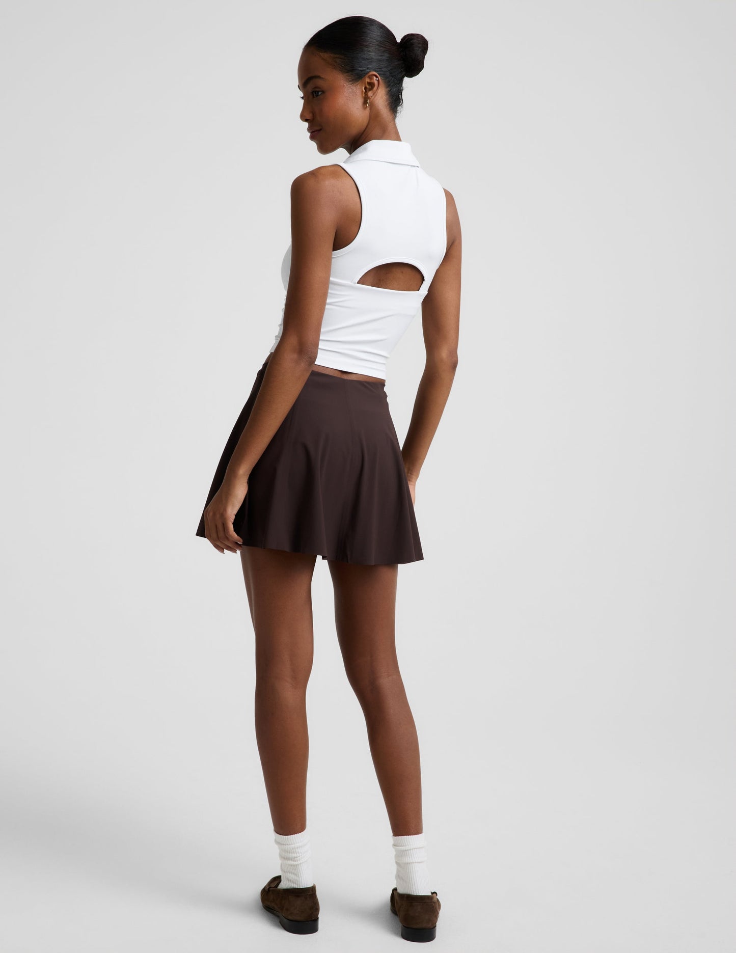 Woman wearing a white polo tank top and brown mini athletic skirt against a plain studio background.