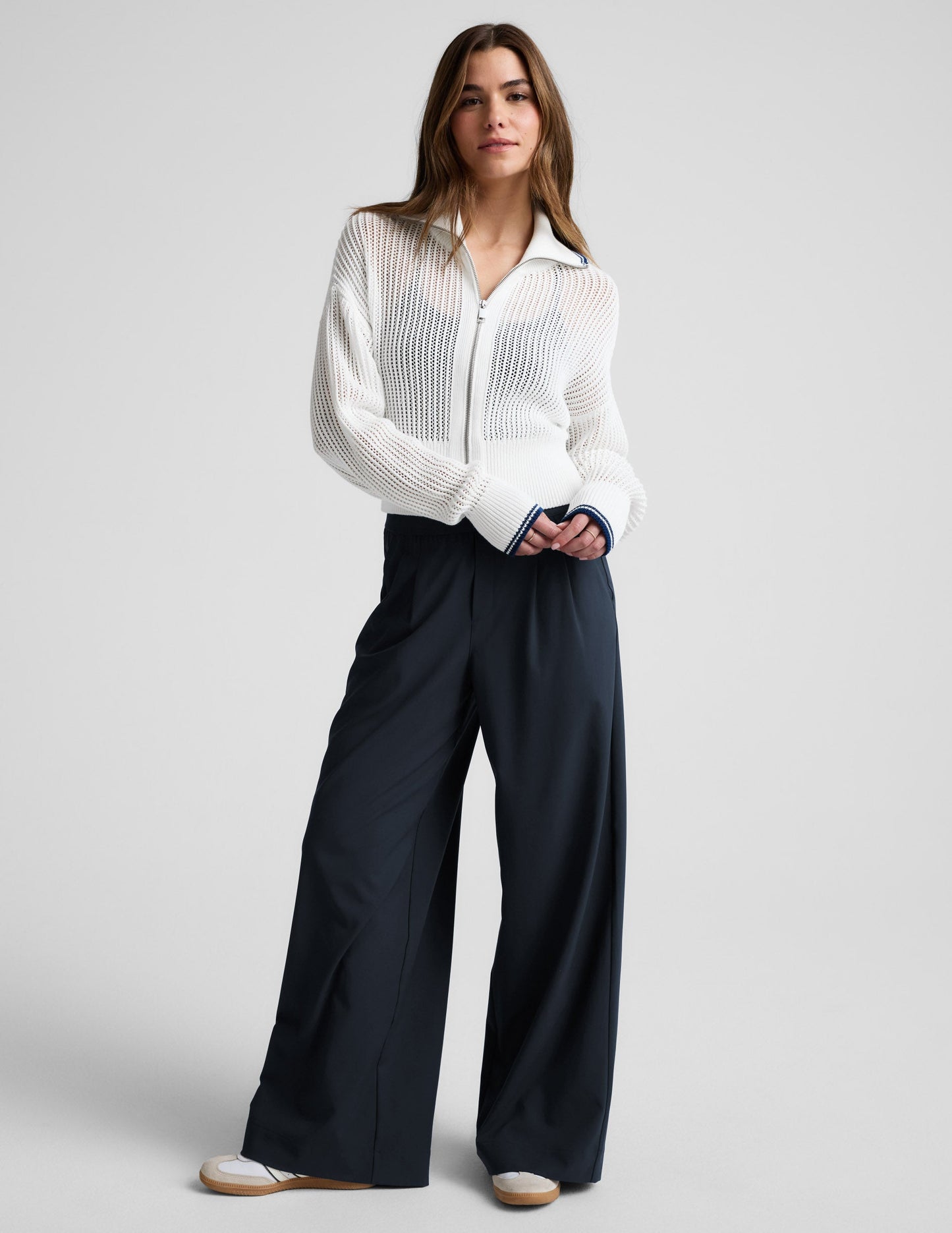 Woman wearing a white zip-up sweater and blue trousers against a plain background.