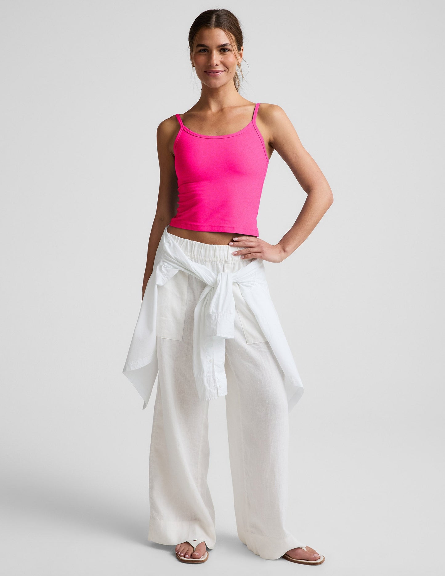 Woman wearing a bright pink waist length tank top and white linen pants against a plain background.