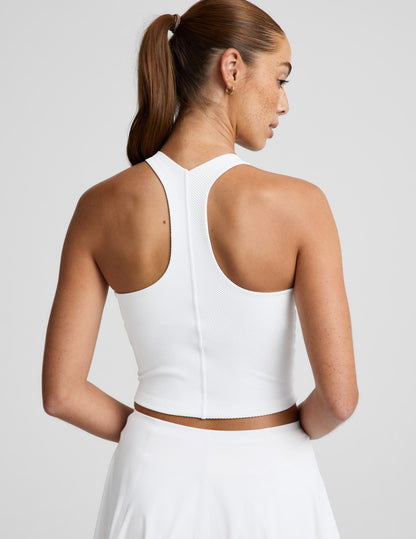 Woman wearing a white ribbed high neck cropped tank and a white skirt against a plain background.