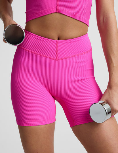 Woman wearing Pink Impulse Ribbed V-Waist 5" Biker Short™ paired with a matching pink top while holding silver dumbbells.