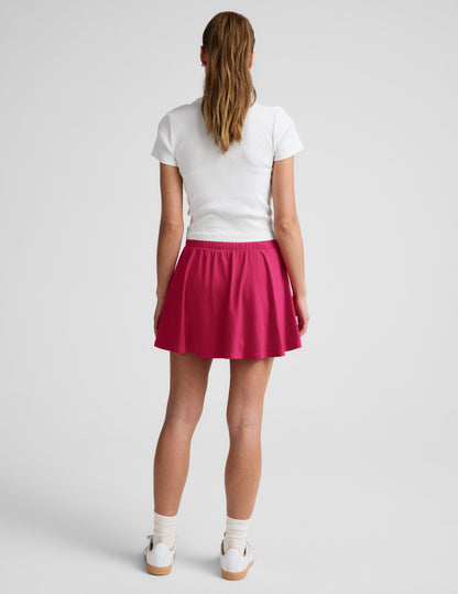 Woman wearing Fuchsia Sport Active Drawcord Mini Skirt, rear view, paired with a white short-sleeve shirt and sneakers.