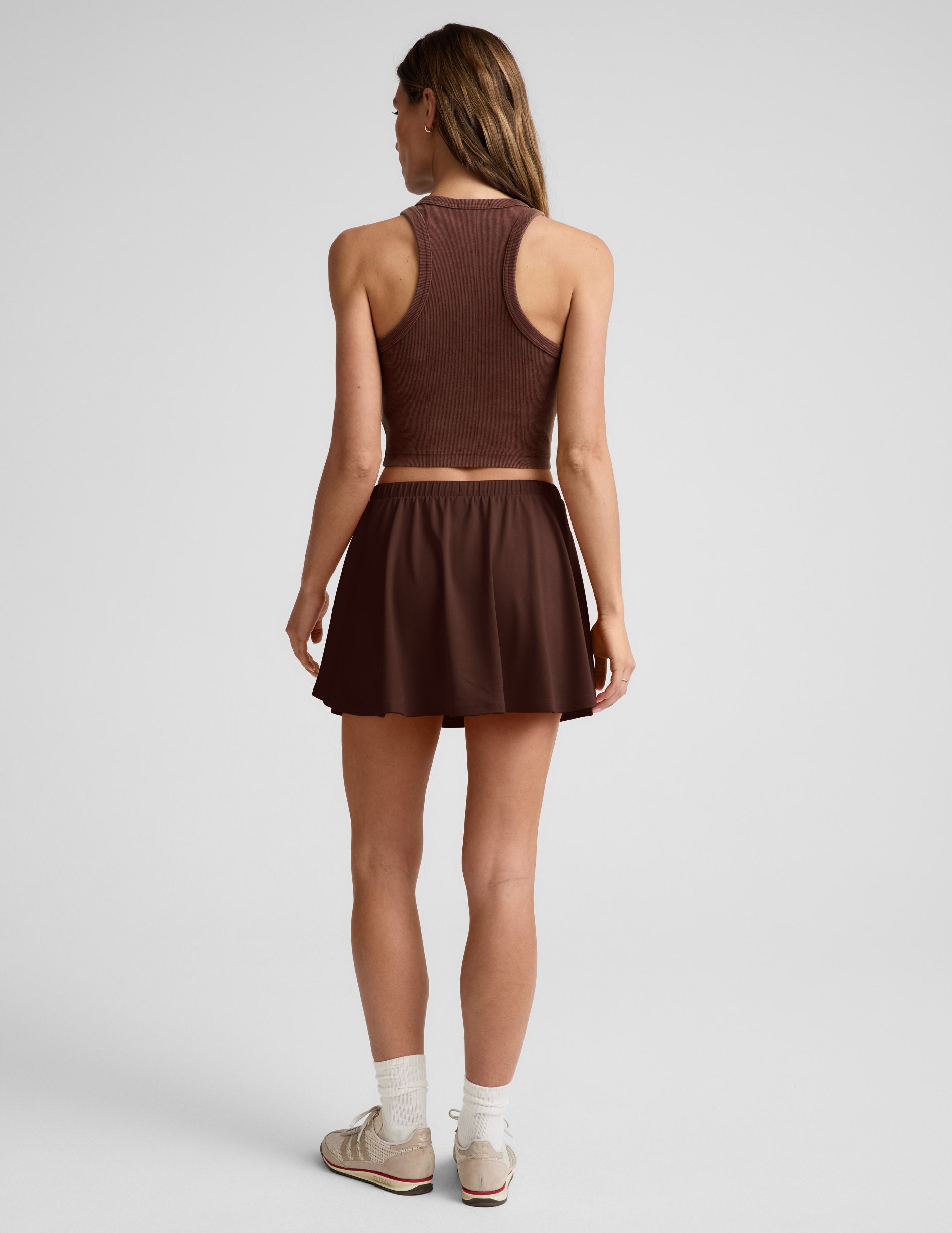 Woman wearing Brown Sport Active Drawcord Mini Skirt from a rear view, paired with a matching tank top and white socks.