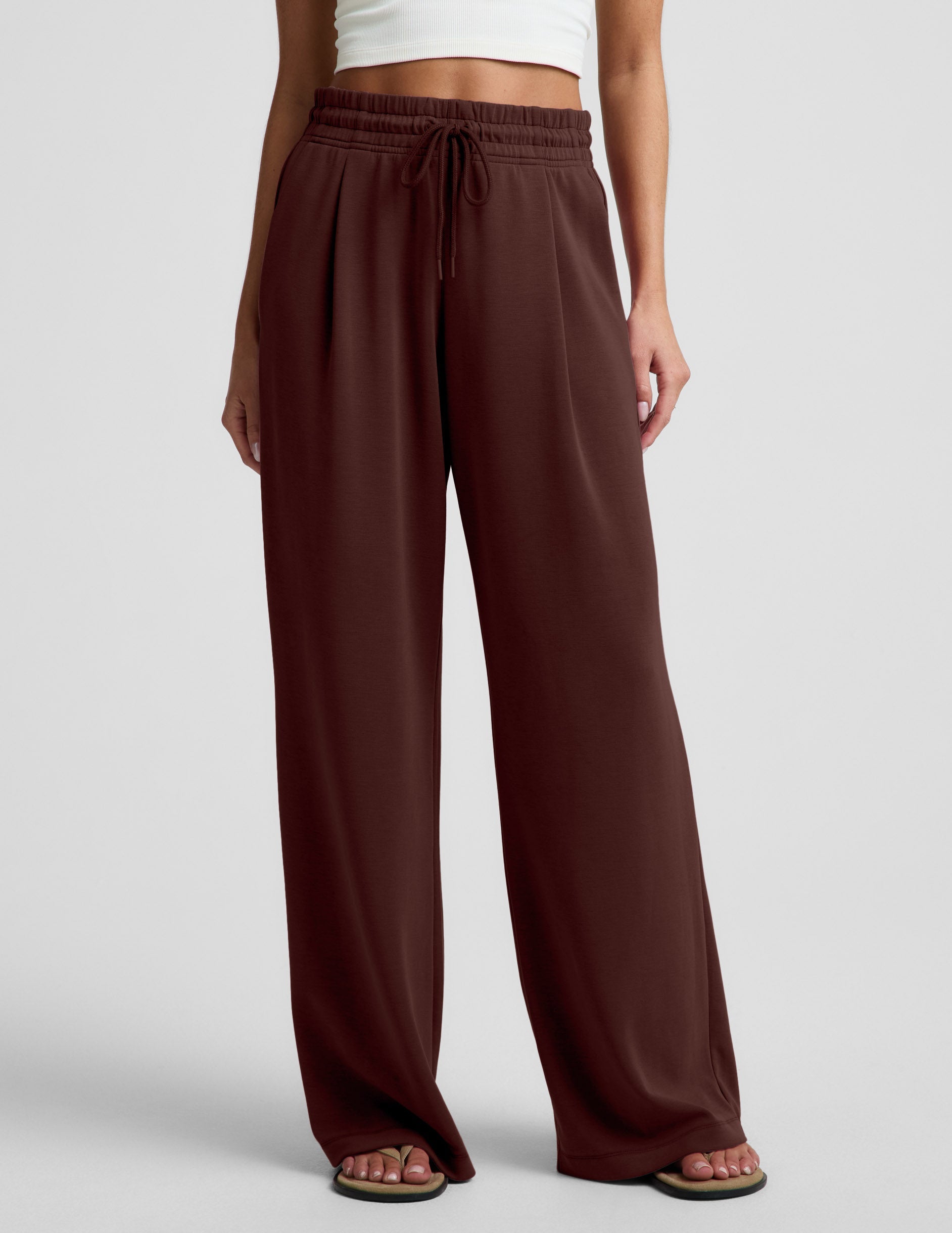 Woman wearing a white cropped tank and brown wide leg pants against a plain background.