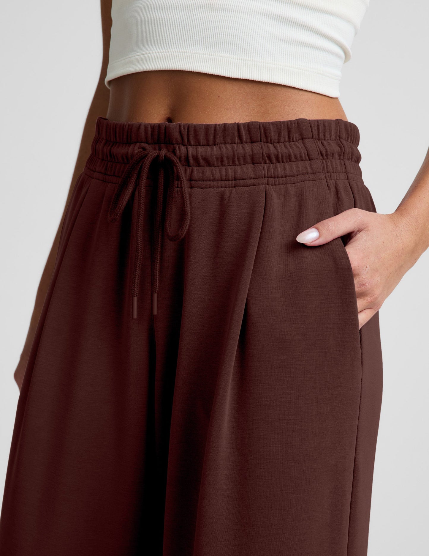 Woman wearing a white cropped tank and brown wide leg pants against a plain background.