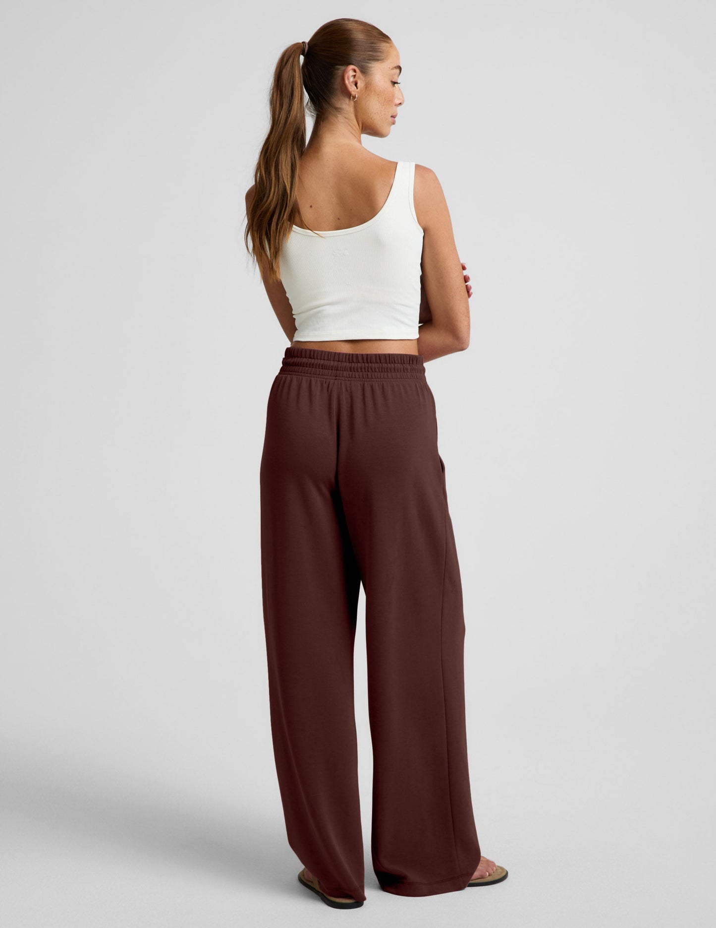 Woman wearing a white cropped tank and brown wide leg pants against a plain background.