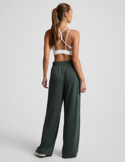 Woman wearing dark green Luxe Sandwash Double Knit Wide Leg Pant from a rear view, styled with a white sports bra.