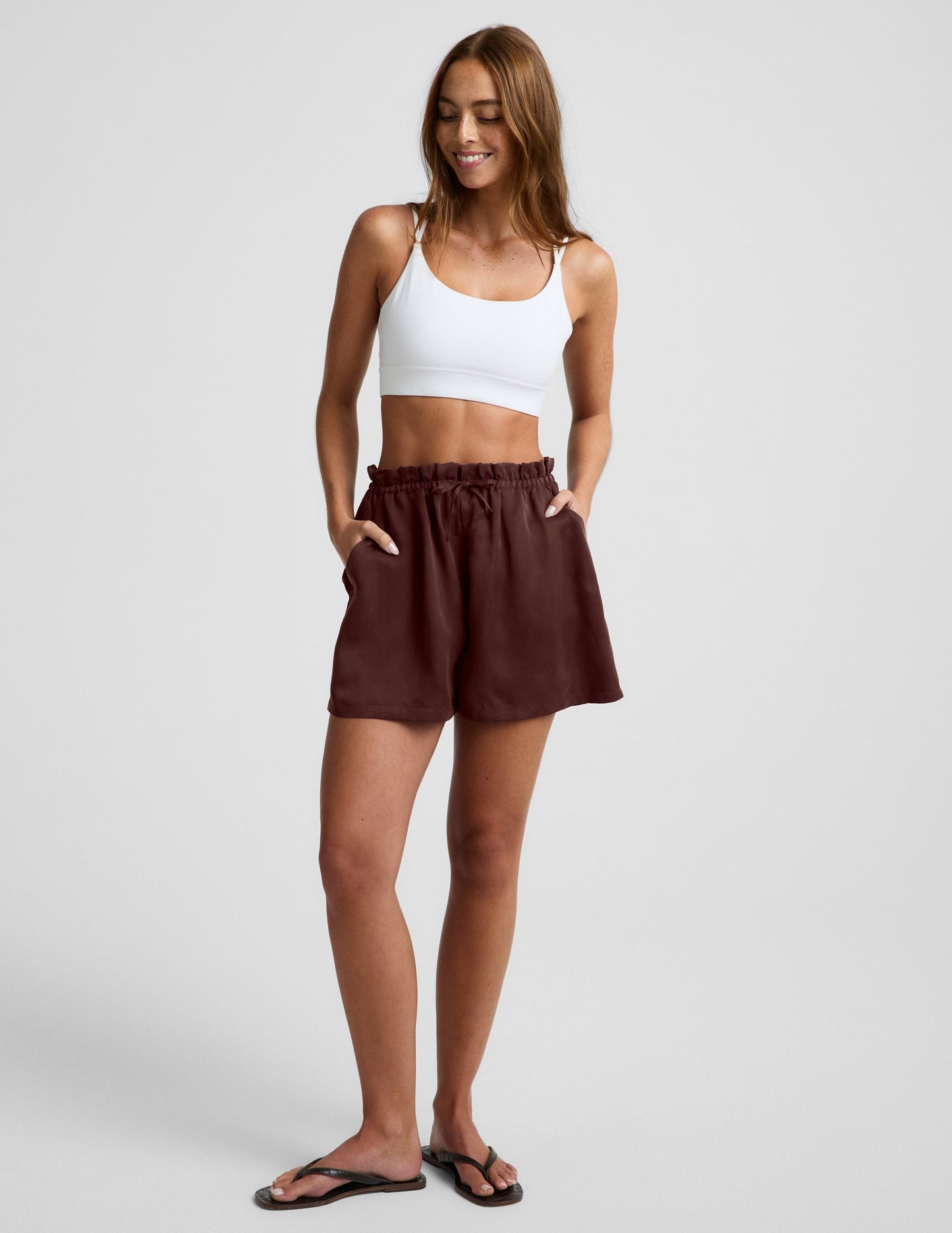 Woman wearing Brown SunKissed Cupro Drawstring Short™ with a flare silhouette, paired with a white sports bra and flip-flops.