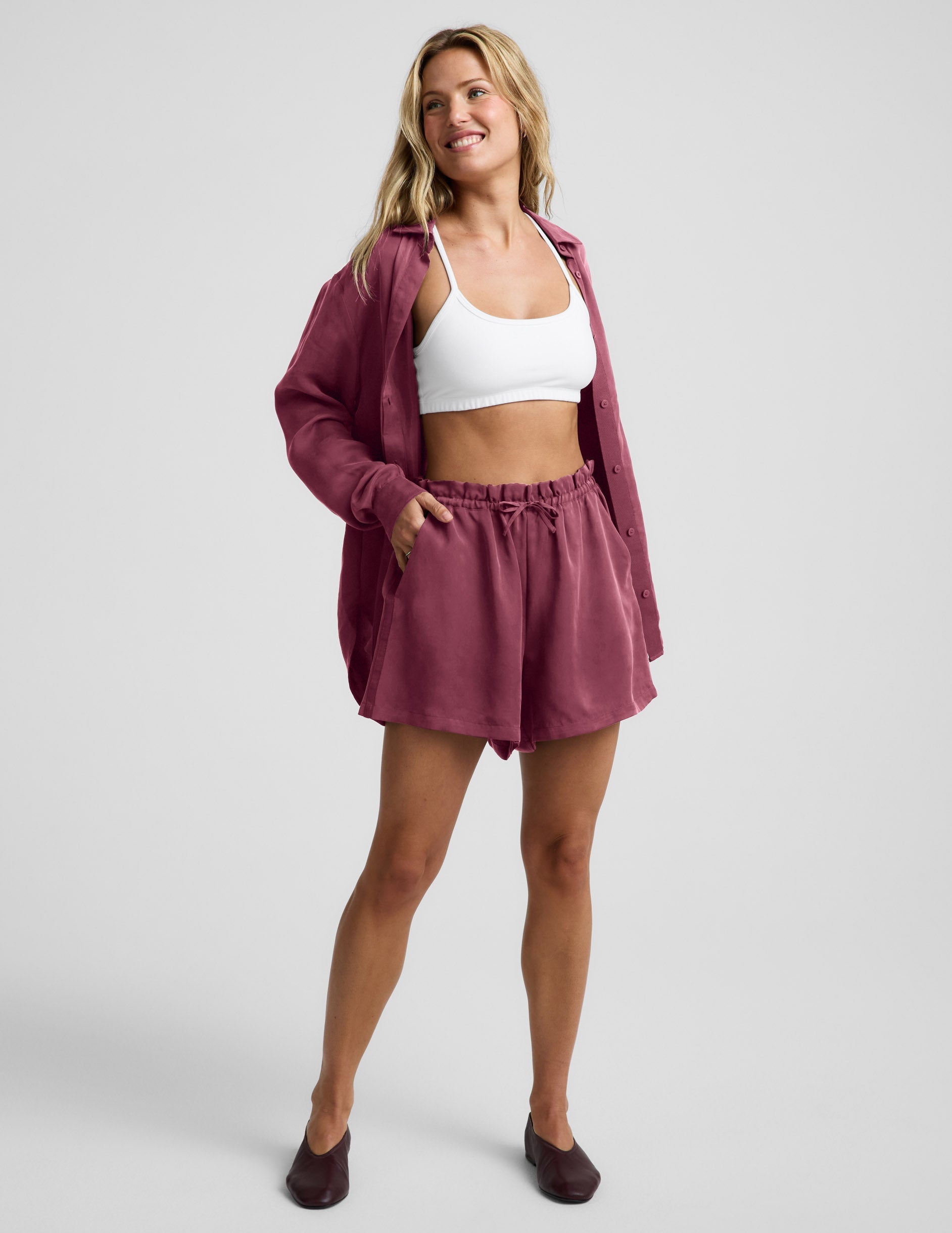 Woman wearing Plum SunKissed™ Cupro Drawstring Short paired with a white sports bra and open matching button-down shirt.