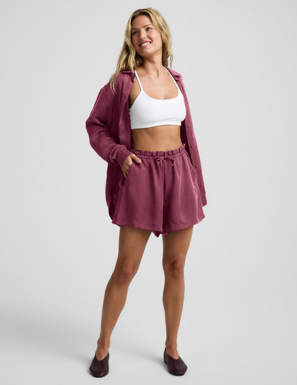 Woman wearing Plum SunKissed™ Cupro Drawstring Short paired with a white sports bra and open matching button-down shirt.