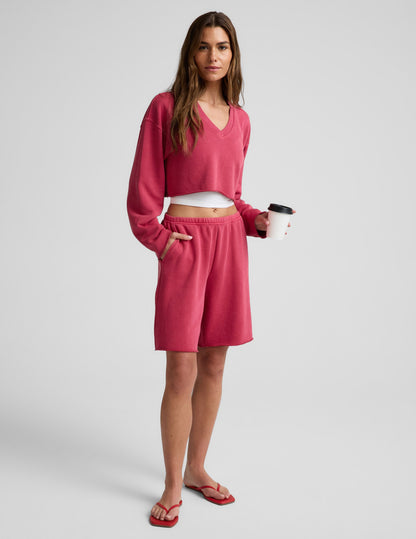 Woman wearing a red cropped long sleeve v-neck sweatshirt and shorts against a plain background.