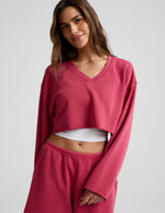 Woman wearing a red cropped long sleeve v-neck sweatshirt and shorts against a plain background.