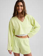 Woman wearing a bright yellow cropped v-neck sweatshirt and sweat shorts against a plain background.