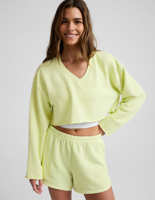 Woman wearing a bright yellow cropped v-neck sweatshirt and sweat shorts against a plain background.