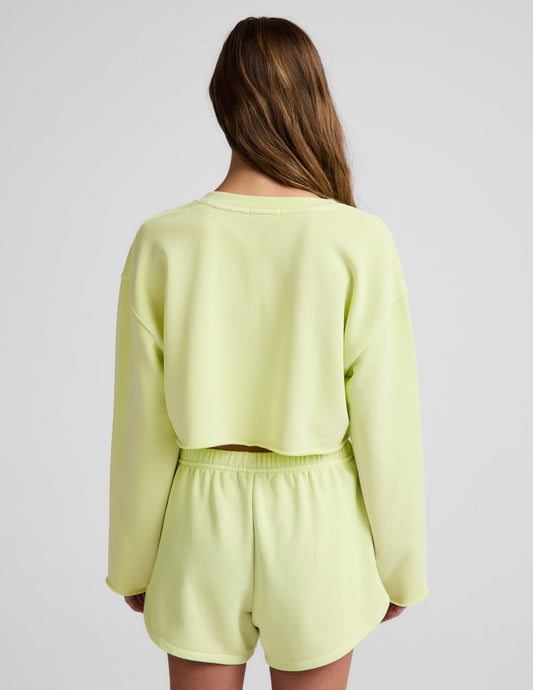 Woman wearing a bright yellow cropped v-neck sweatshirt and sweat shorts against a plain background.