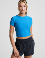 Woman wearing a bright blue lightweight cropped t-shirt and blue athletic shorts against a plain background.