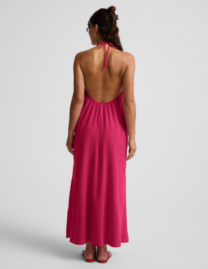 Woman wearing a red halter maxi dress with an open back against a light gray background.