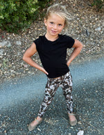 A young girl posing outdoors on a gravel path, wearing a black short-sleeve t-shirt and leopard-print leggings.