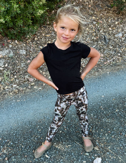 A young girl posing outdoors on a gravel path, wearing a black short-sleeve t-shirt and leopard-print leggings.