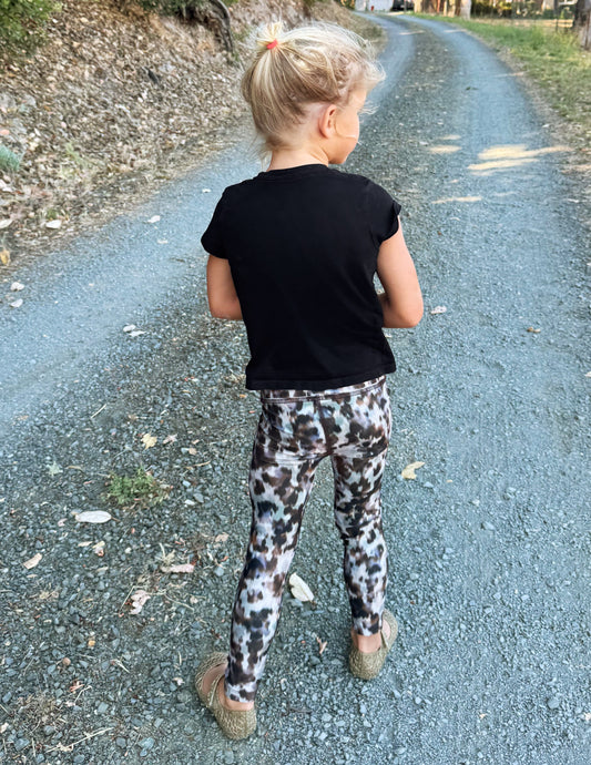 A young girl posing outdoors on a gravel path, wearing a black short-sleeve t-shirt and leopard-print leggings.