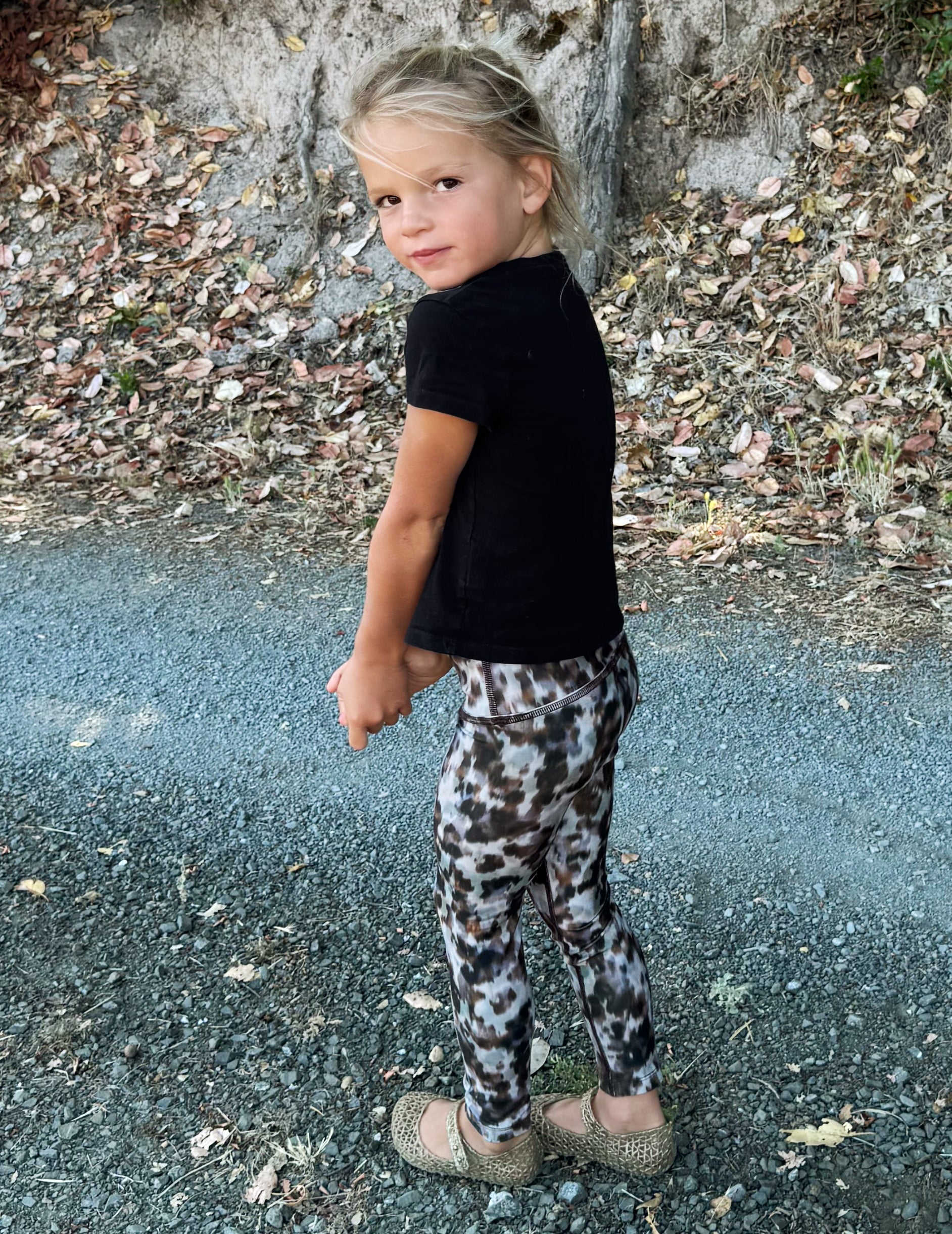 A young girl posing outdoors on a gravel path, wearing a black short-sleeve t-shirt and leopard-print leggings.