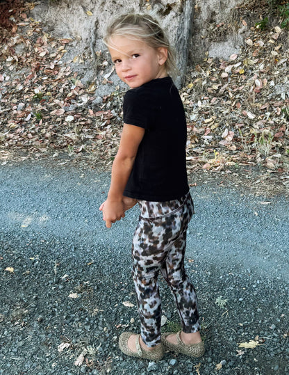A young girl posing outdoors on a gravel path, wearing a black short-sleeve t-shirt and leopard-print leggings.