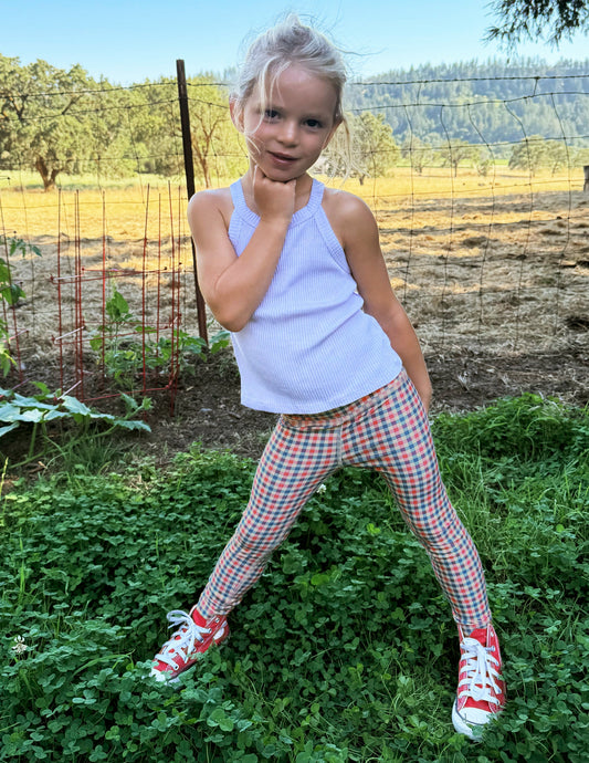 Toddler wearing a white tank top and gingham printed leggings standing on grass.