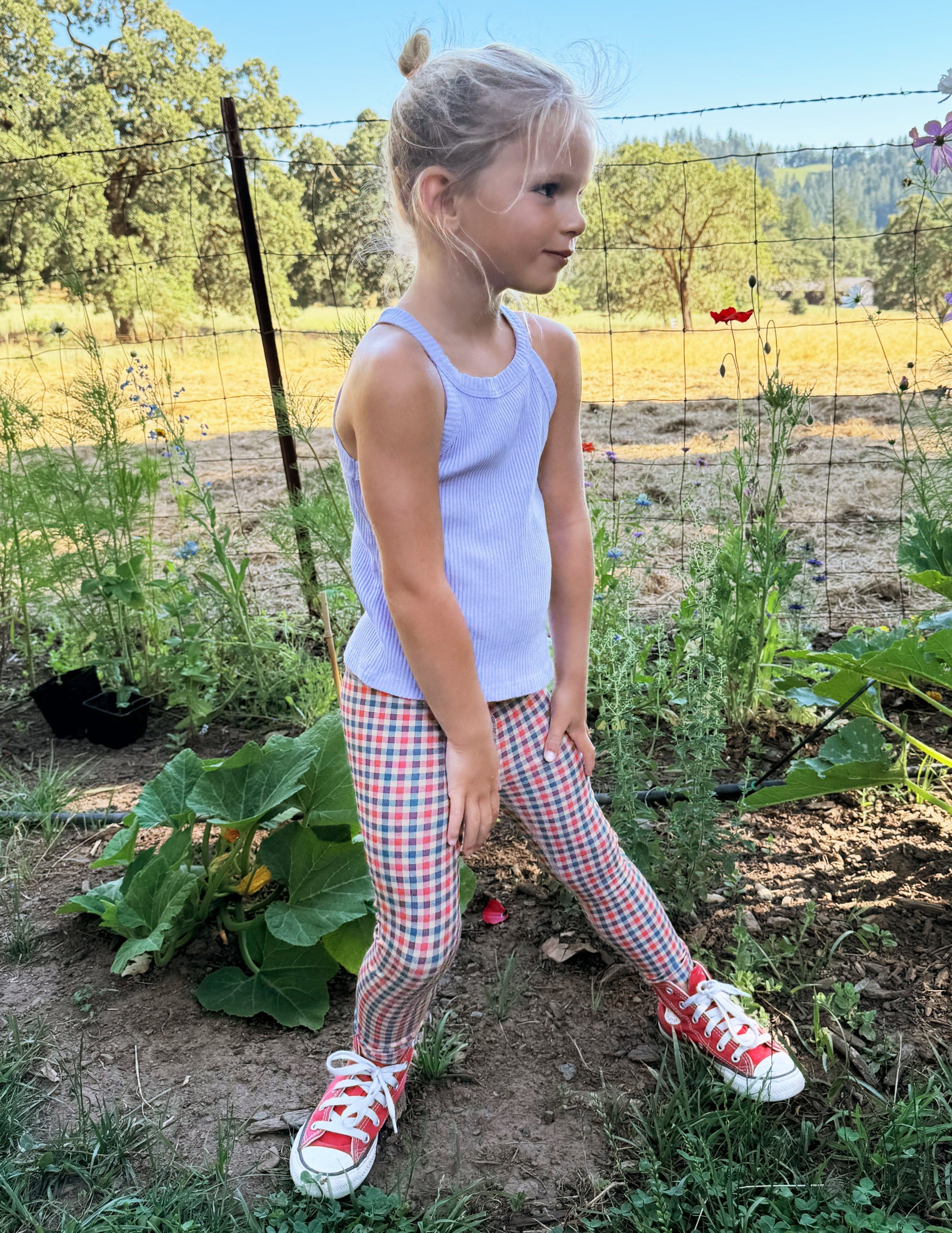 Toddler wearing a white tank top and gingham printed leggings standing on grass.