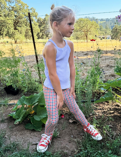 Toddler wearing a white tank top and gingham printed leggings standing on grass.