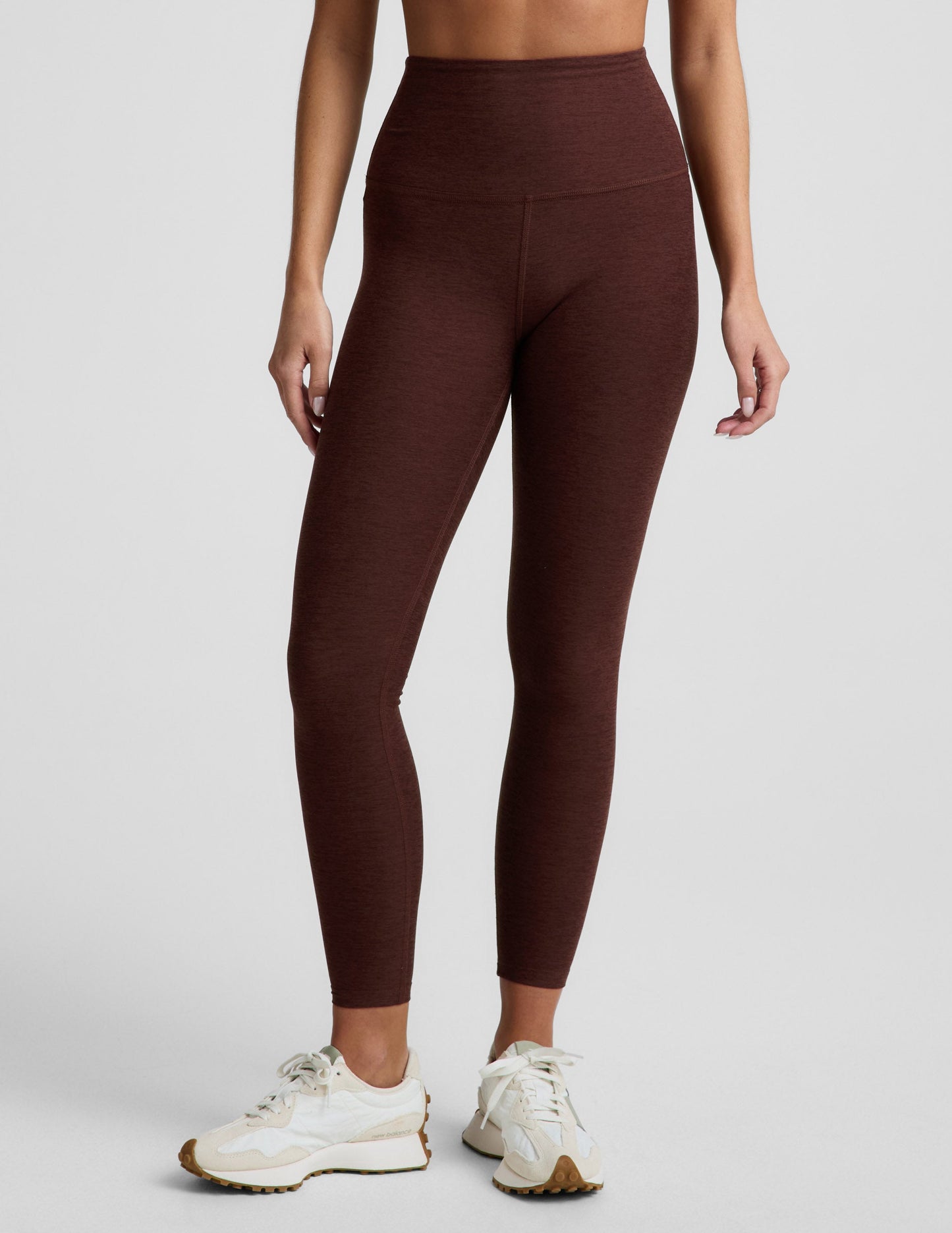 Woman wearing a brown sports bra and brown high-waisted midi length leggings against a plain background.