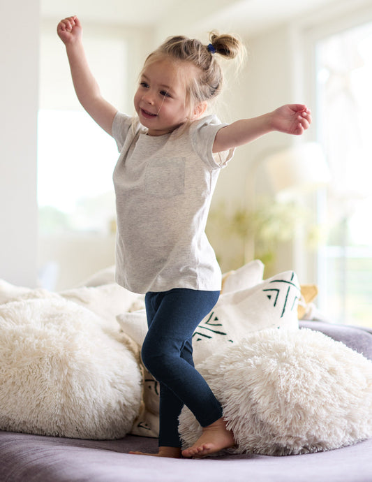 Girl wearing Navy Spacedye™ Toddler Legging jumping on a sofa, paired with a light grey pocket t-shirt.